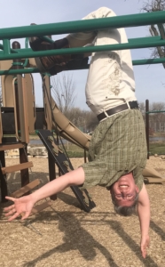 Sasha hanging from their knees, upside down on a playground climbing thing. They are smiling at the camera, expressing joy.